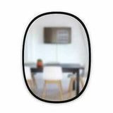 The Hub Mirror Oval - Black from the Umbra range features a stylish rubber rim, complemented by a sleek table and chairs in the background.