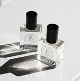 Two clear square glass bottles of SASS Perfume Oil with black caps, photographed in soft natural light with shadows and reflections on a white surface.

