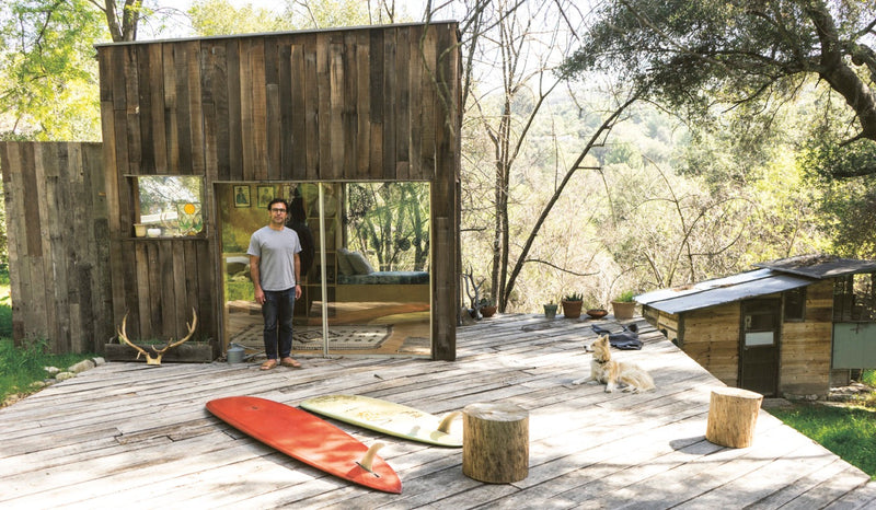 A man standing on a wooden deck with surfboards outside of his Surf Shacks: An Eclectic Compilation Of Creative Surfers Homes From Coast To Coast and Overseas book.