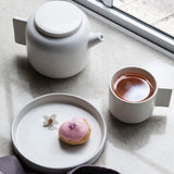 A white Tab Plate - Various Sizes teapot and a pink donut on a window sill, both dishwasher safe. Brand: Zakkia