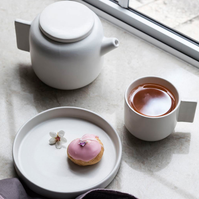 A white Tab Plate - Various Sizes teapot and a pink donut on a window sill, both dishwasher safe. Brand: Zakkia