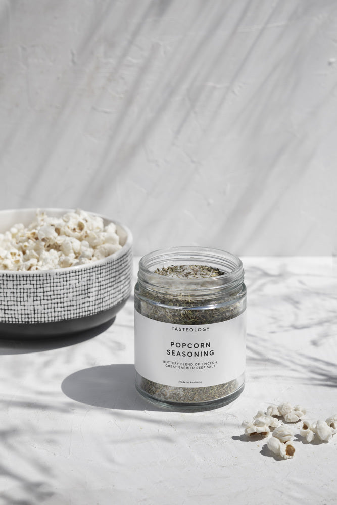 Tasteology Popcorn Seasoning jar styled outdoors with a bowl of popcorn and sunlight casting soft shadows.