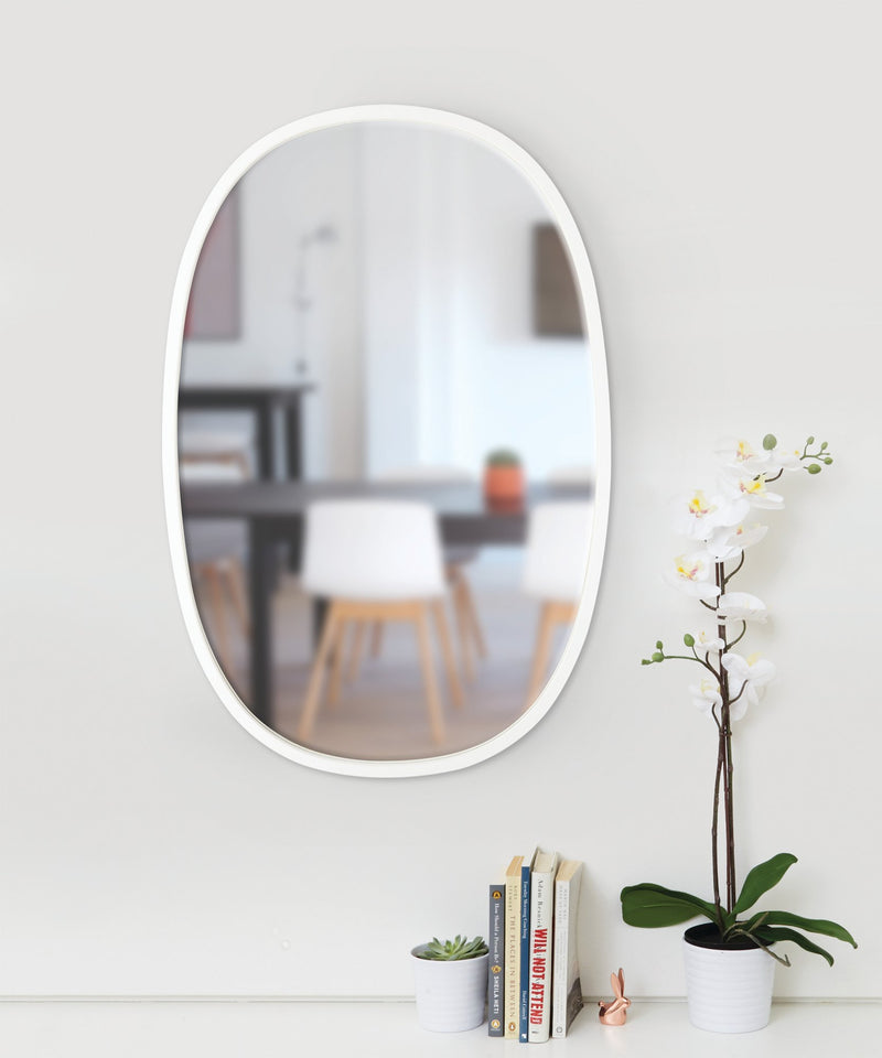 An Umbra Hub Mirror Oval 61 x 91 Grey hanging on a wall next to a plant and books.