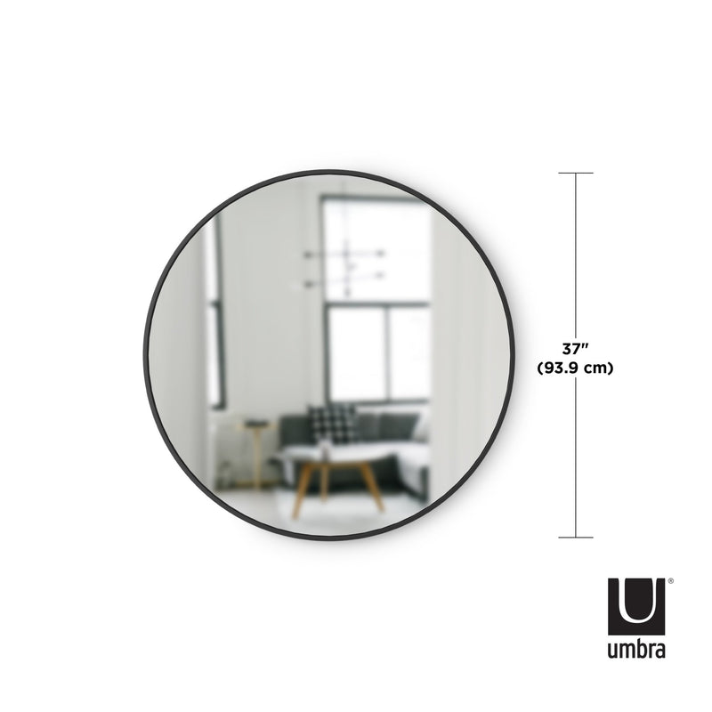 A modern round mirror, the Umbra HUB MIRROR 45CM BLACK, is shown in front of a living room.