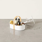 A modern white caddy with a curved wood handle sits on a concrete floor, filled with neatly arranged skincare bottles and sponges. 