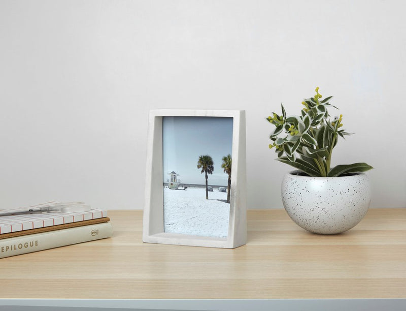 A modern Edge Marble Frame, part of the Umbra range, on a desk next to a plant and a book.