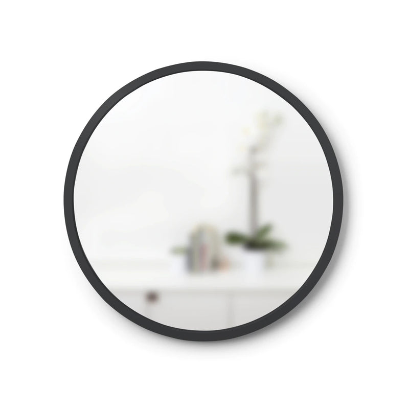 A modern round mirror, the HUB MIRROR 45CM BLACK by Umbra, placed on a sleek white surface with a vibrant plant elegantly displayed within it.