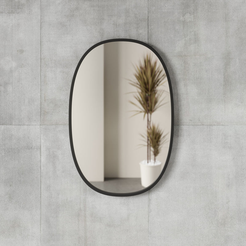 The Hub Mirror Oval - Black from the Umbra range is mounted on a concrete wall and features a sleek black design. Accentuating its modern look, the mirror is framed with a stylish rubber rim.