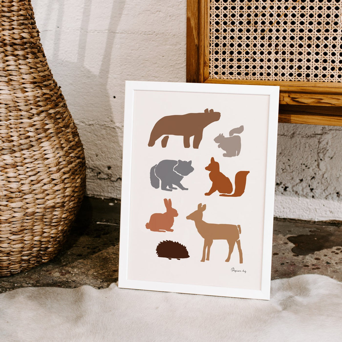 Animal Woodland print in a white frame leaning on a rattan chair beside a woven vase