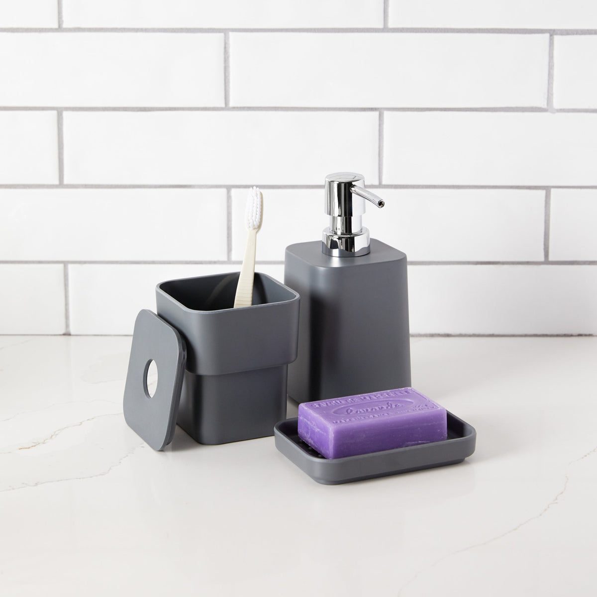 Charcoal SCILLAE canister holding a toothbrush beside a matching soap pump and soap dish on white tiles