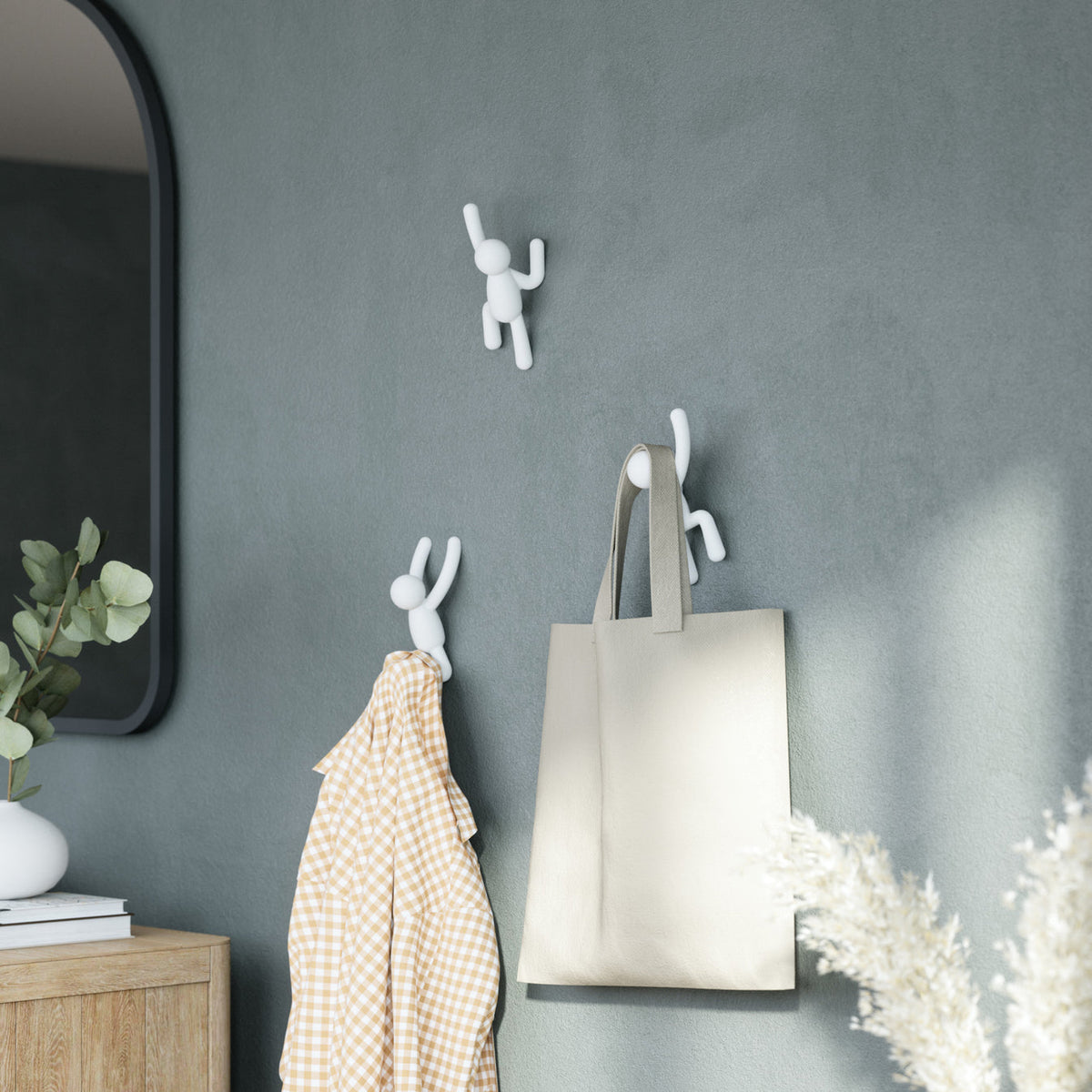 Umbra Buddy hooks in white on a dark grey wall holding a tote bag and a check towel